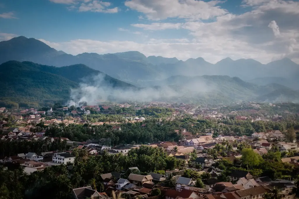 Aerial view of Luang Prabang, Laos, surrounded by lush green mountains, traditional rooftops, palm trees, and misty clouds under a bright blue sky
