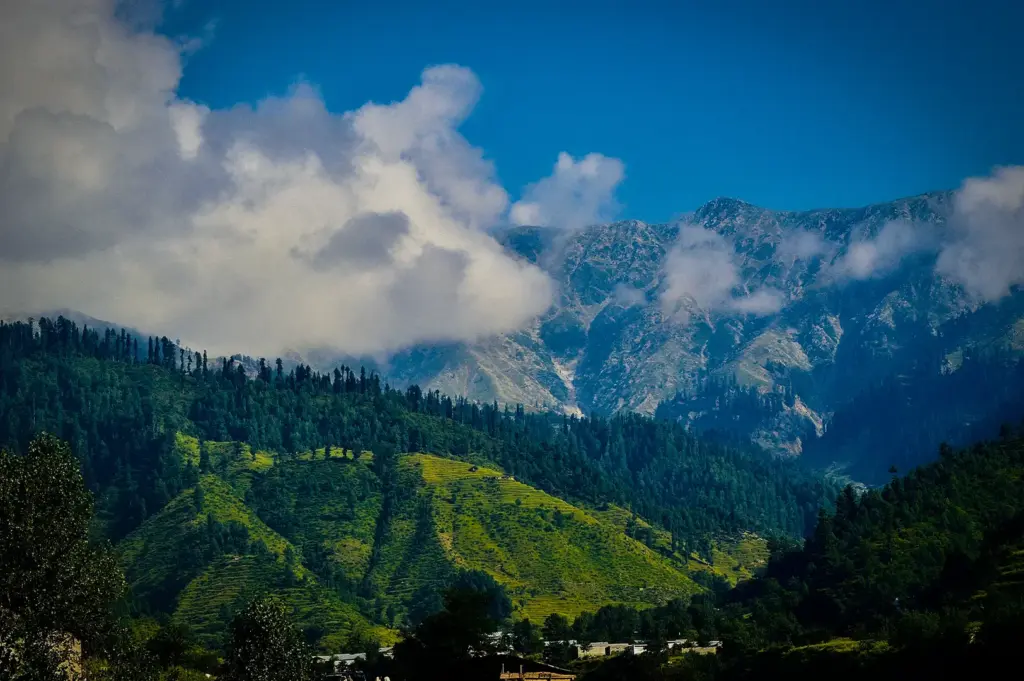 Mountain landscape with lush green hills, dense pine forests, rugged peaks, drifting clouds, and a small settlement in the valley below.