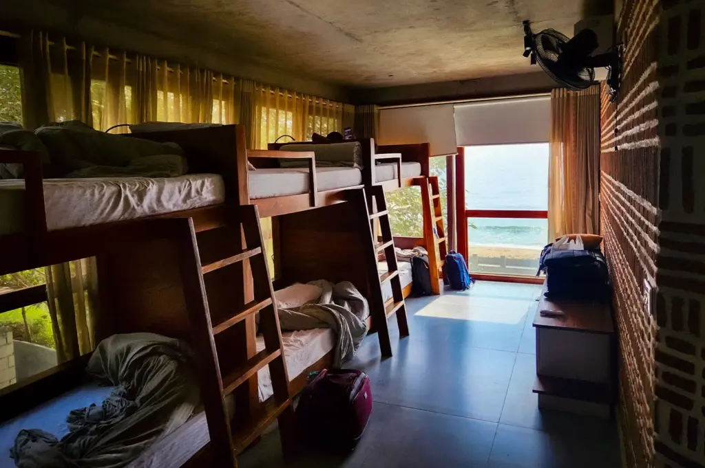 Modern beachfront hostel dorm room with wooden bunk beds, large windows, ocean views, and shared accommodations ideal for budget travelers and backpackers.