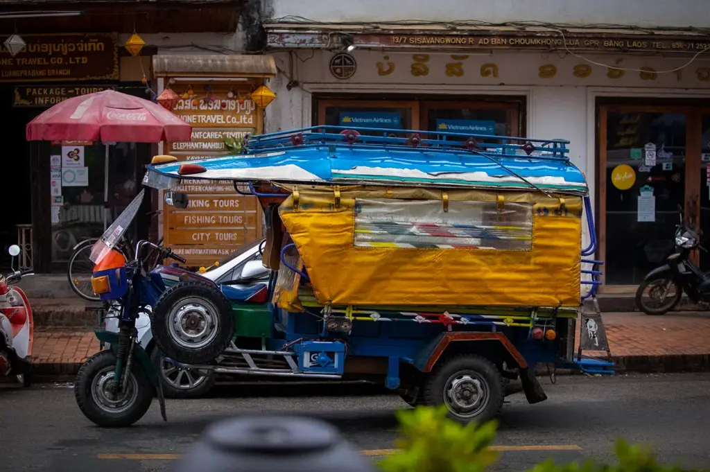Colorful blue and yellow tuk-tuk parked on a street in Luang Prabang, Laos, with traditional buildings, travel shops, and local signs in the background