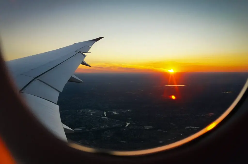 Airplane wing viewed through a passenger window during sunset, with the sun glowing above a city landscape and distant horizon.