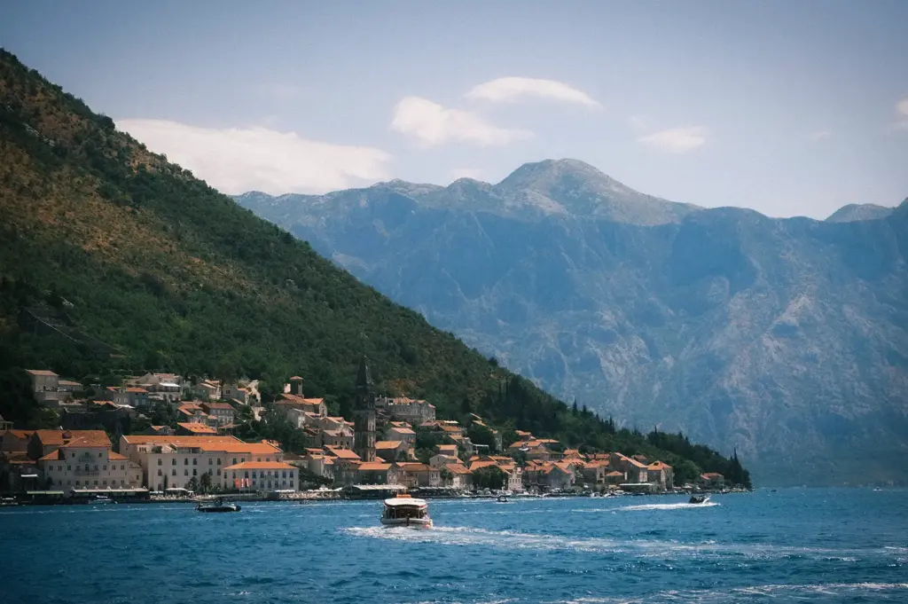Scenic coastal village with terracotta rooftops, boats on blue water, forested hills, and dramatic mountains surrounding a tranquil bay.
