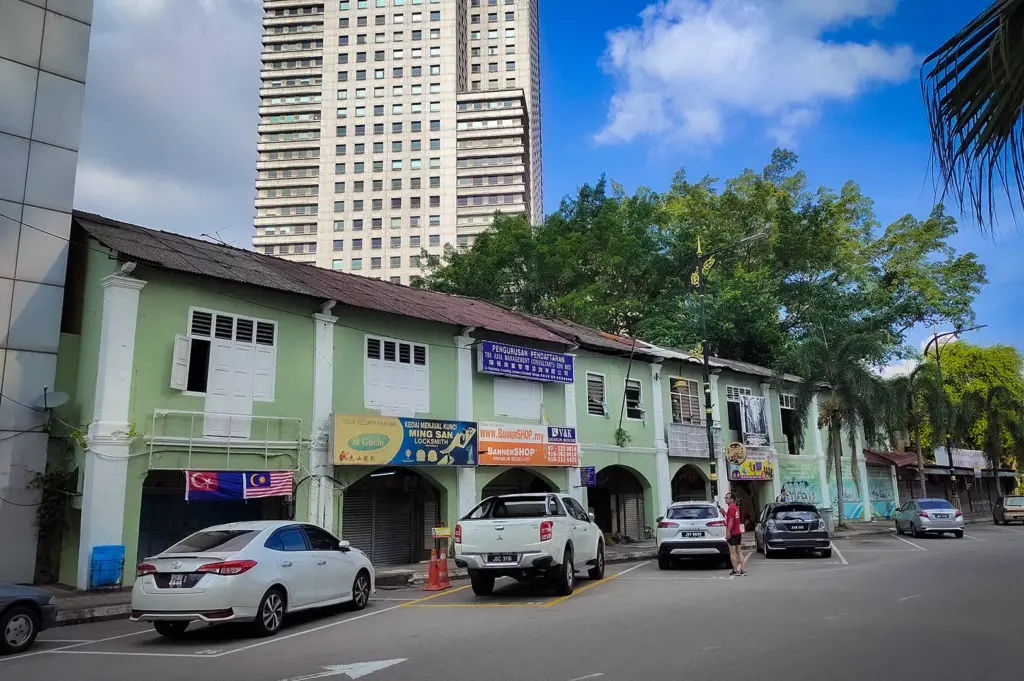 Johor Bahru street with historic shop houses, parked cars, and a modern high-rise building in the background
