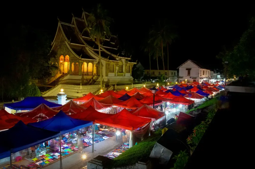 Luang Prabang Night Market in Laos illuminated at night with colorful red and blue stalls beside a traditional Buddhist temple