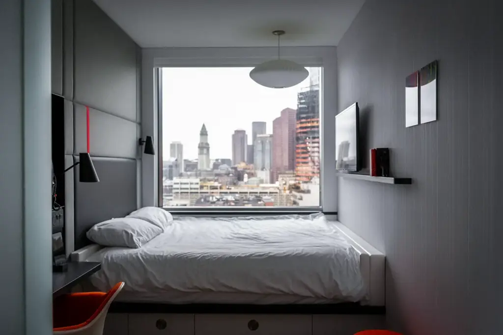 Contemporary hotel room with a large bed, floor-to-ceiling window overlooking the city skyline, minimalist décor, workspace, and modern amenities for urban travelers.