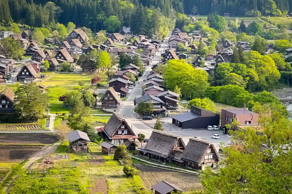 Traditional Japanese village with thatched-roof houses, rice fields, forested mountains, winding roads, and lush green countryside.