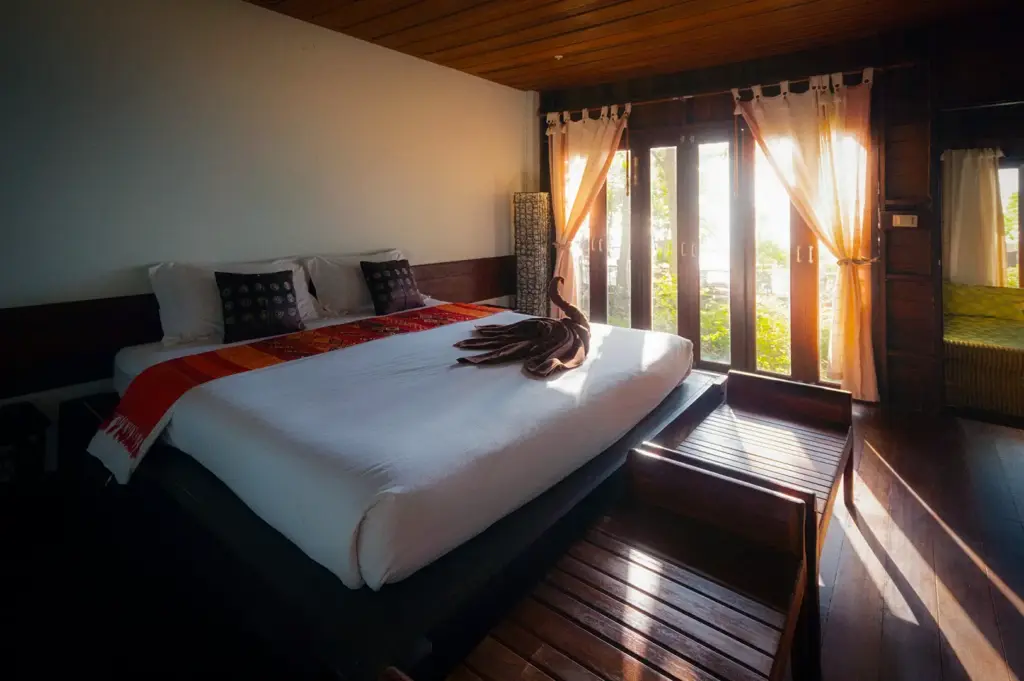 Spacious wooden homestay bedroom with a king-size bed, large glass doors, garden views, warm natural light, and rustic interiors for a relaxing tropical stay.