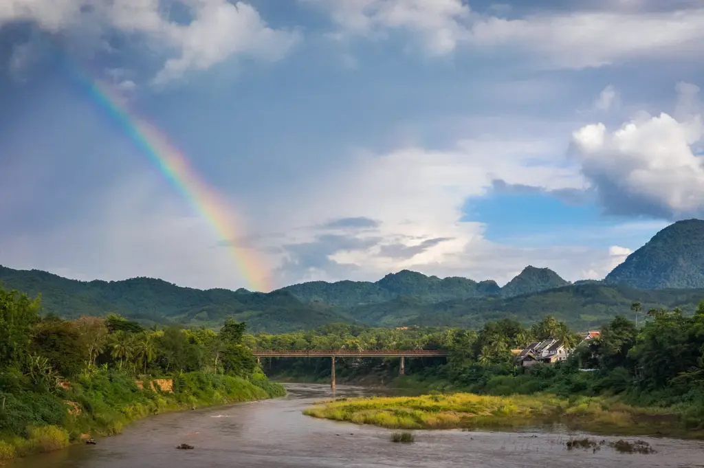 Nam Khan River in Luang Prabang, Laos, flowing beneath an old bridge with lush mountains, tropical vegetation, and a rainbow beneath a cloudy sky