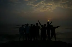 Silhouettes standing together on a rocky shoreline facing the ocean against a dramatic sunset sky.