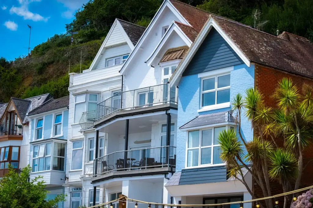 Row of colorful multi-story coastal holiday homes and cottages built along a lush green hillside, featuring light blue siding, gabled roofs, and palm trees in the foreground