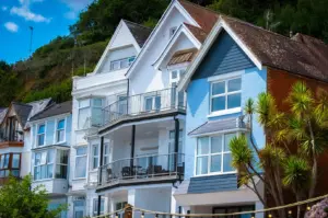 Row of colorful multi-story coastal holiday homes and cottages built along a lush green hillside, featuring light blue siding, gabled roofs, and palm trees in the foreground