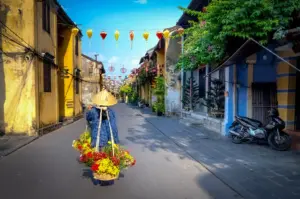 Street vendor wearing a traditional conical hat carrying colorful flowers through the yellow-walled streets of Hoi An Ancient Town in Vietnam