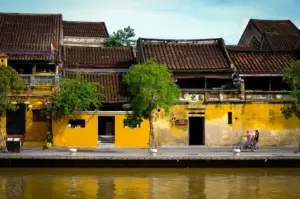 Hoi An, Vietnam ancient town riverside with historic yellow buildings, traditional tiled roofs, lush trees, and colorful reflections along the river.