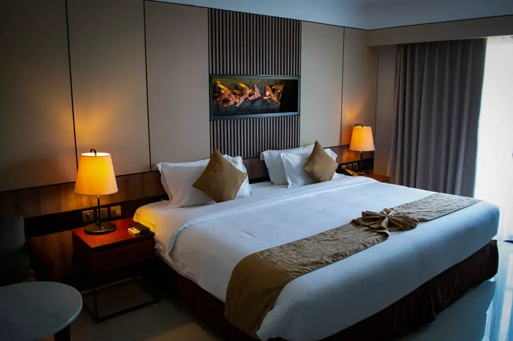 Modern hotel room featuring a king bed with white linens, brown bed runner, bedside lamps, and decorative wall paneling.