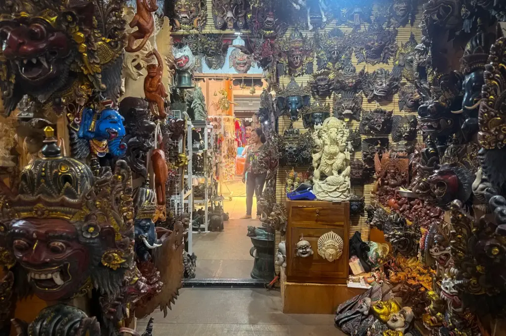 A bustling shop interior at the Ubud Art Market in Ubud, Bali, Indonesia, packed floor-to-ceiling with colorful traditional wooden masks, carved statues, and local handicrafts.