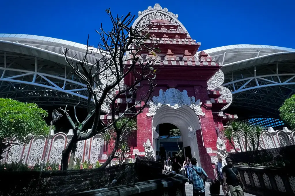 An exterior view of I Gusti Ngurah Rai International Airport in Bali, Indonesia, featuring traditional Balinese red brick gateway architecture under a clear blue sky.