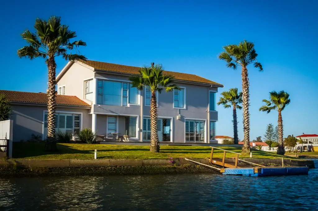 Modern two-story waterfront holiday home rental featuring a lush green lawn, tall palm trees, and a private wooden boat dock on a canal under a clear blue sky