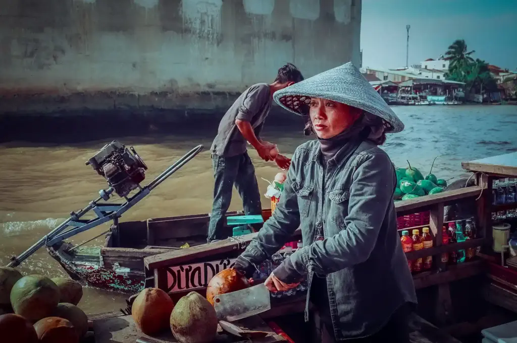 Vietnamese vendor wearing a conical hat chopping fresh coconuts on a wooden boat at a river market in Cam Thanh, Vietnam