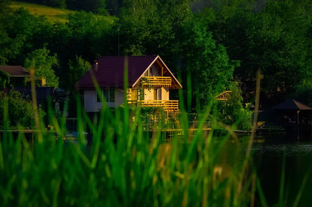 Scenic view through tall green grass of a lakeside holiday cabin featuring wooden balconies, nestled against a dense forest and calm water