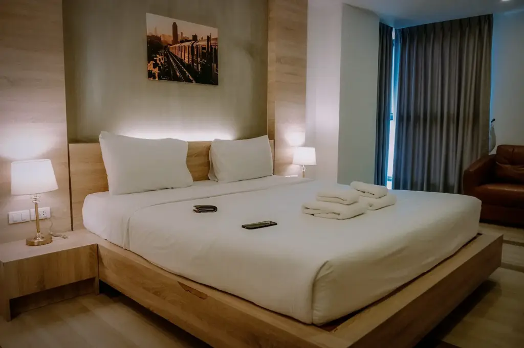 Cozy modern hotel bedroom featuring a wooden platform bed with crisp white linens, neatly folded towels, bedside lamps, and a framed wall artwork.