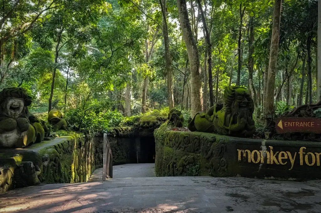 The shaded stone entranceway to the Sacred Monkey Forest Sanctuary in Ubud, Bali, Indonesia, featuring moss-covered mythological stone statues and lush trees.