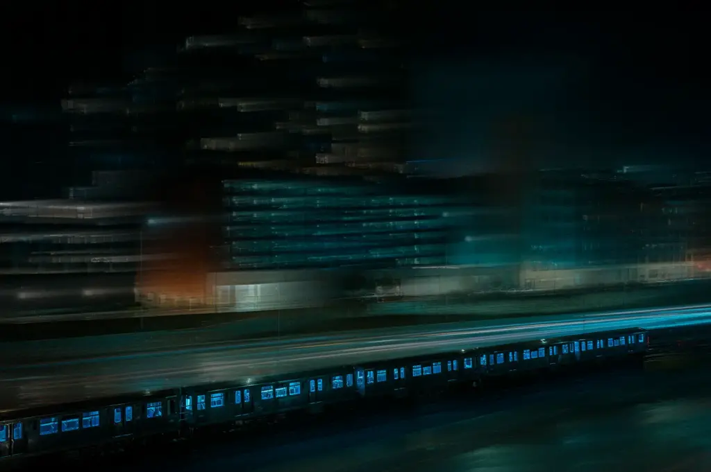 Nighttime cityscape with a passenger train, illuminated buildings, and motion-blurred light trails.