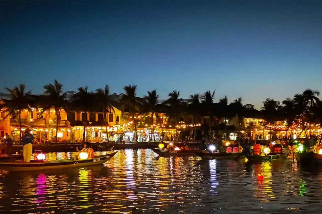 Thu Bon River in Hoi An, Vietnam, at night with illuminated boats, colorful lanterns, riverside buildings, and reflections across the water.