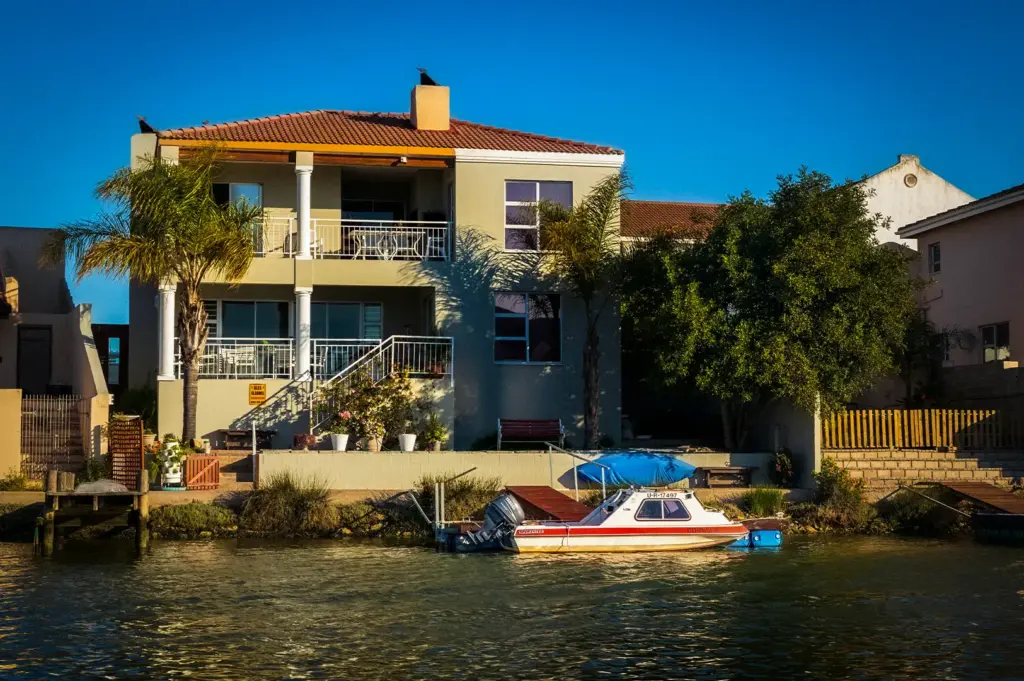 Two-story waterfront holiday villa featuring upper and lower covered balconies, palm trees, and a small motorboat moored at a private dock on a canal