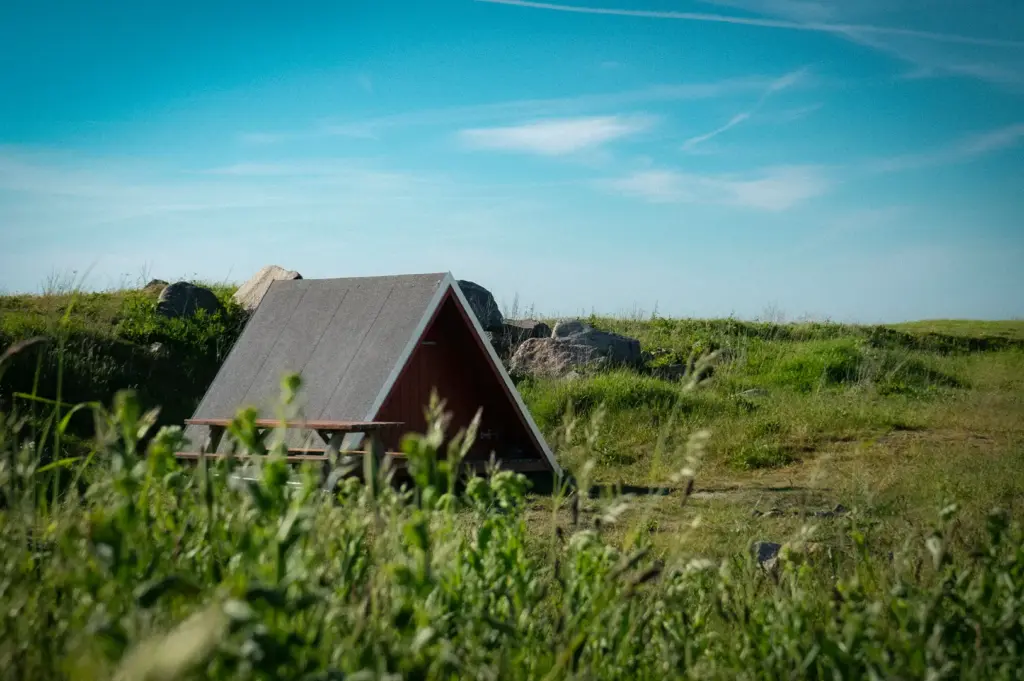 Small A-frame wooden cabin with a steep roof and outdoor wooden picnic table set in a grassy open meadow under a bright blue sky