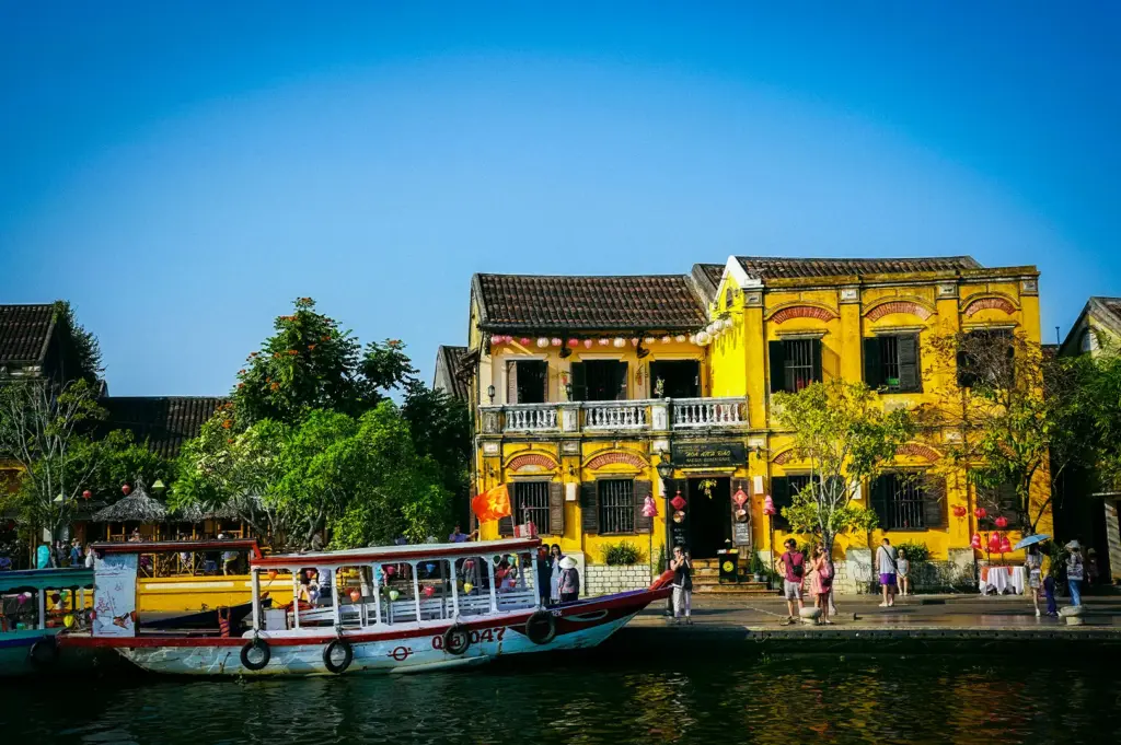 Hoi An, Vietnam, with historic yellow buildings, colorful boats, lanterns, and the riverside waterfront under a clear blue sky.