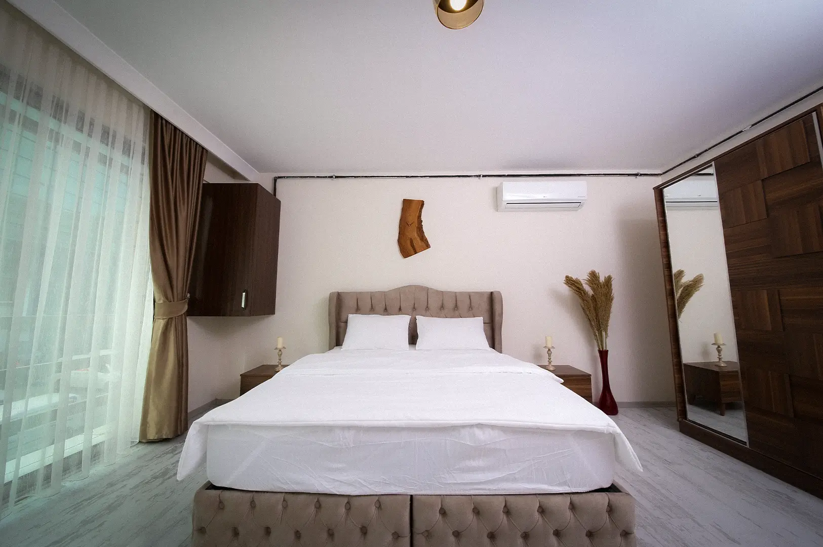 Bright modern hotel bedroom featuring a upholstered king bed with white bedding, wooden wardrobe, and large window with sheer curtains.