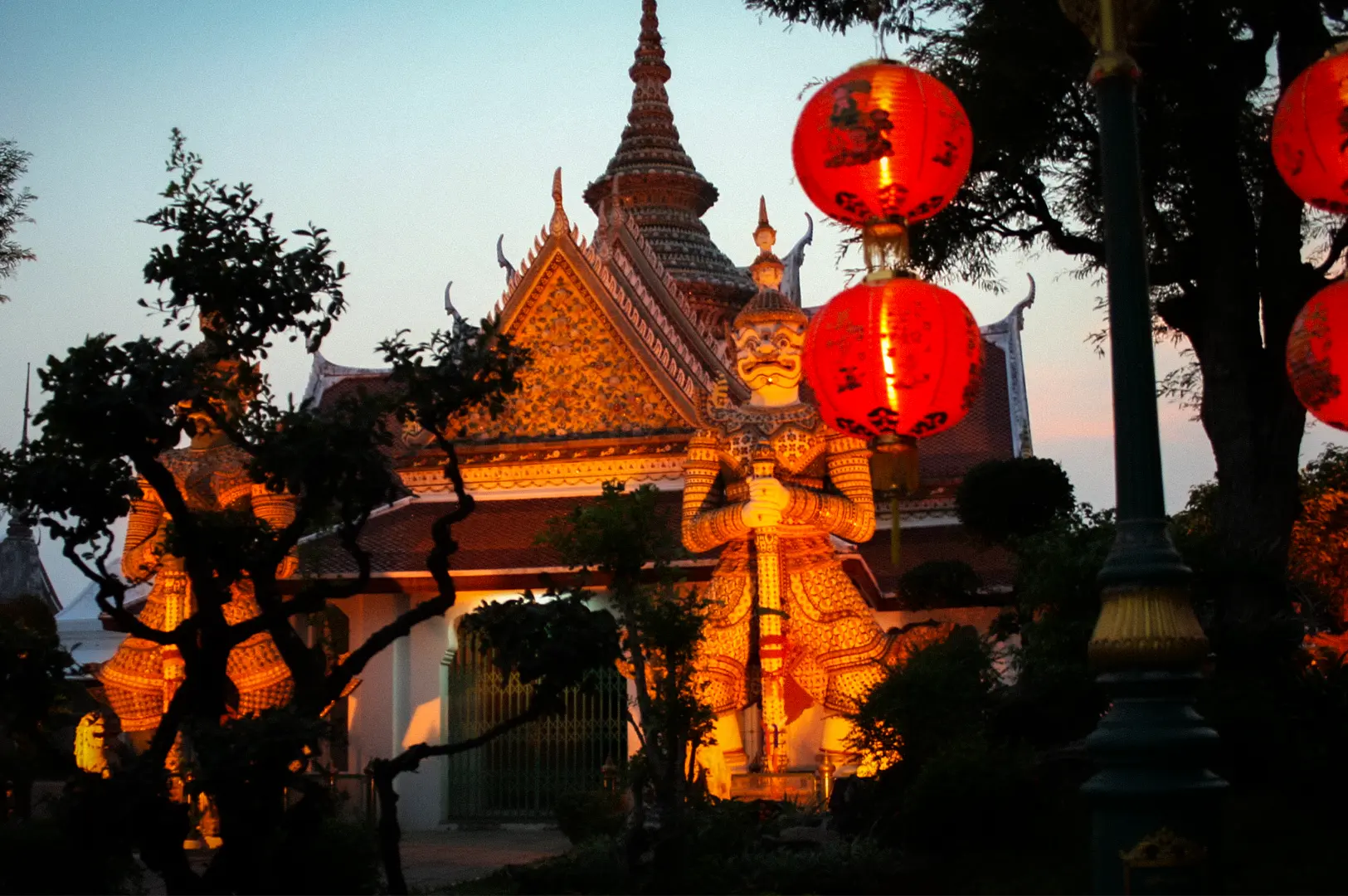 Bangkok temple at dusk with illuminated Thai architecture, guardian statue, and red lanterns in the evening light
