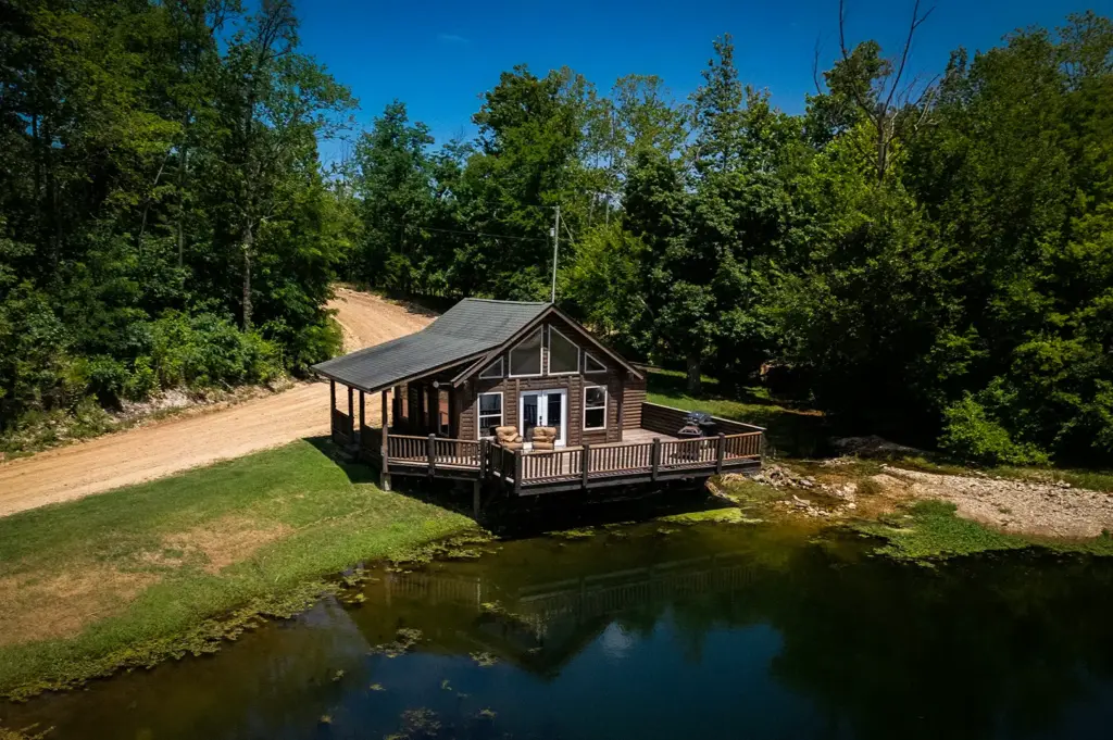 High-angle aerial view of a secluded wooden log cabin with a large wrap-around deck overlooking a quiet pond near a dirt road and dense green forest