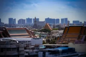 An elevated cityscape view of Bangkok, Thailand, featuring traditional ornate temple roofs in the foreground, historic temple spires in the mid-ground, and modern skyscrapers rising on the skyline under a bright sky.