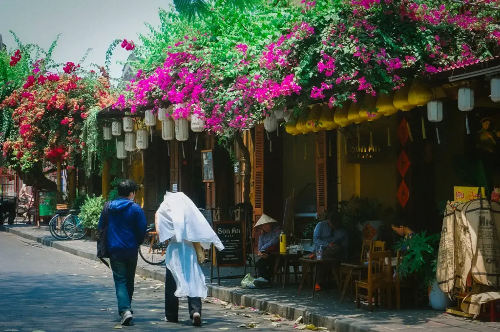 Pedestrians walking past Son An Cafe along a street lined with vibrant pink bougainvillea flowers and hanging yellow lanterns in Hoi An Ancient Town