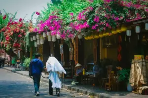 Pedestrians walking past Son An Cafe along a street lined with vibrant pink bougainvillea flowers and hanging yellow lanterns in Hoi An Ancient Town