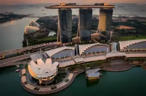 An aerial view of Marina Bay Sands and the ArtScience Museum in Singapore at sunset, showcasing the iconic three-tower hotel structure, rooftop Skypark, and surrounding waterfront bay area.