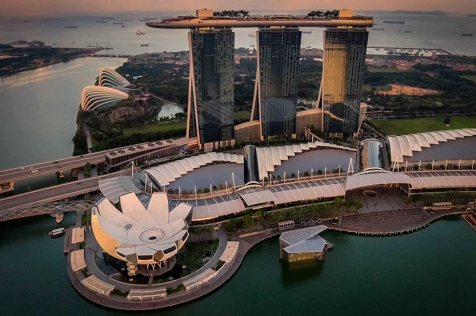 An aerial view of Marina Bay Sands and the ArtScience Museum in Singapore at sunset, showcasing the iconic three-tower hotel structure, rooftop Skypark, and surrounding waterfront bay area.