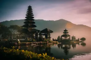 A scenic landscape view of Ubud, Bali, featuring traditional multi-tiered temple pagodas sitting by the tranquil water of a lake under a soft morning sky.