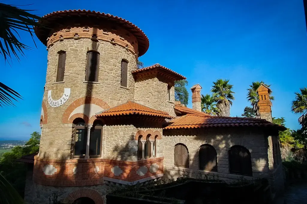 Exterior stone facade and tower of the historic Villa Sassetti in Sintra, Portugal, featuring terracotta tile roofing, arched windows, and a sundial set against a blue sky