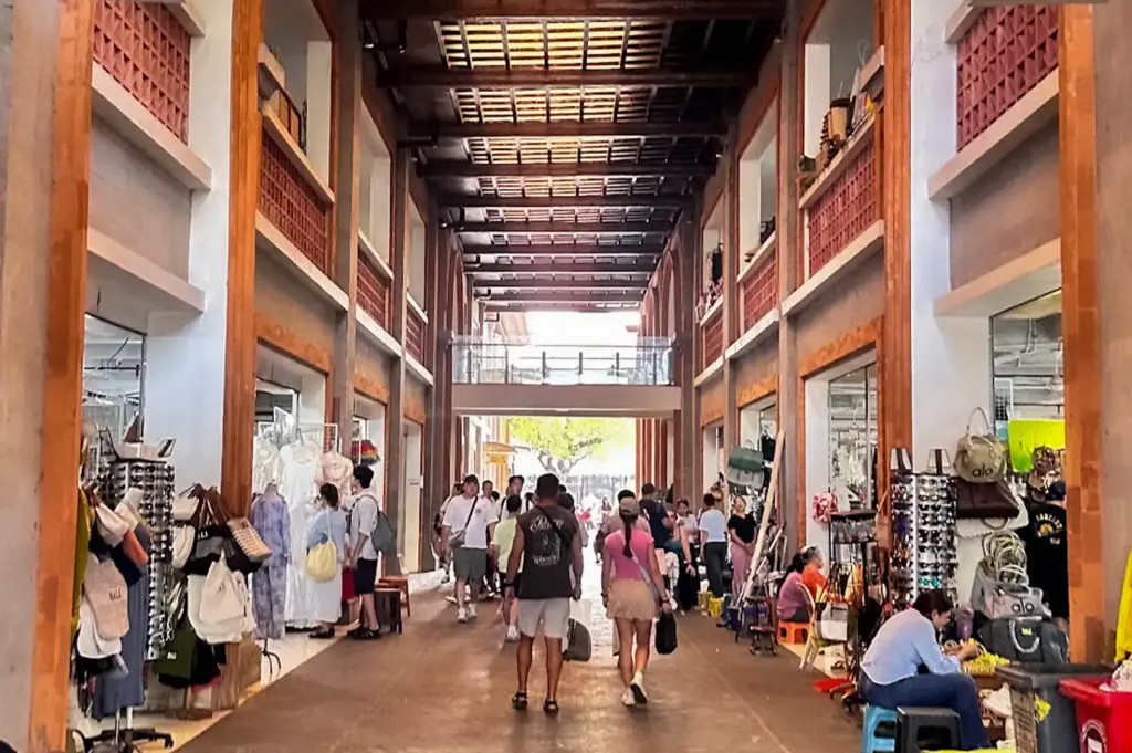 An indoor corridor view of the Ubud Art Market in Bali, featuring high skylit ceilings, traditional pillars, local vendors, and visitors walking through the marketplace.