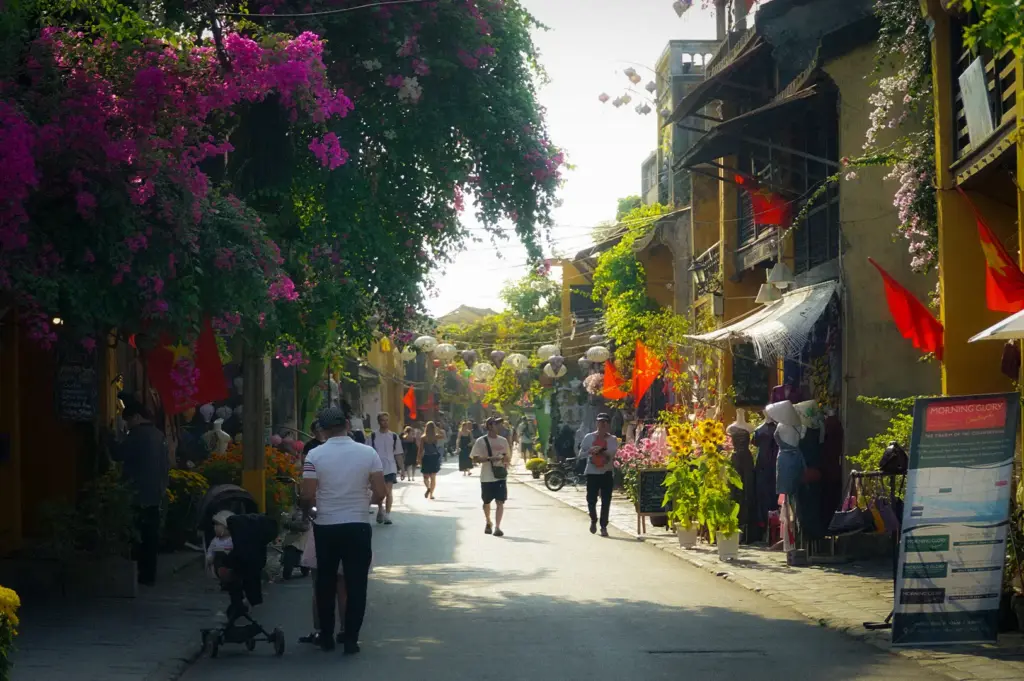 Pedestrians strolling down a sunny street in Hoi An Ancient Town lined with yellow shophouses, vibrant pink bougainvillea flowers, Vietnamese flags, and a Morning Glory restaurant display standee
