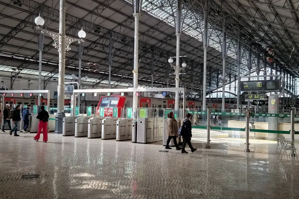Interior concourse and ticket gates of Rossio Railway Station in Lisbon, Portugal, featuring high iron-truss ceilings, pillars, and travelers walking on a cobblestone floor