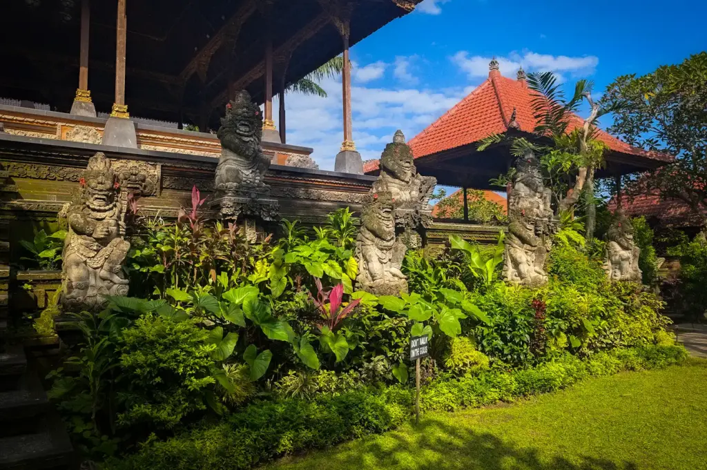 An outdoor view of Puri Saren Agung Ubud ( Puri Saren Ubud ) in Bali, featuring traditional Balinese stone guardian statues amidst lush green foliage against a bright blue sky.