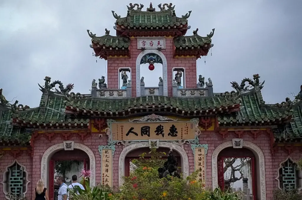 Ornate triple-arched entrance gate of the Fujian Assembly Hall in Hoi An, featuring green tiled roofs, Chinese calligraphic signage, and potted plants in the foreground courtyard