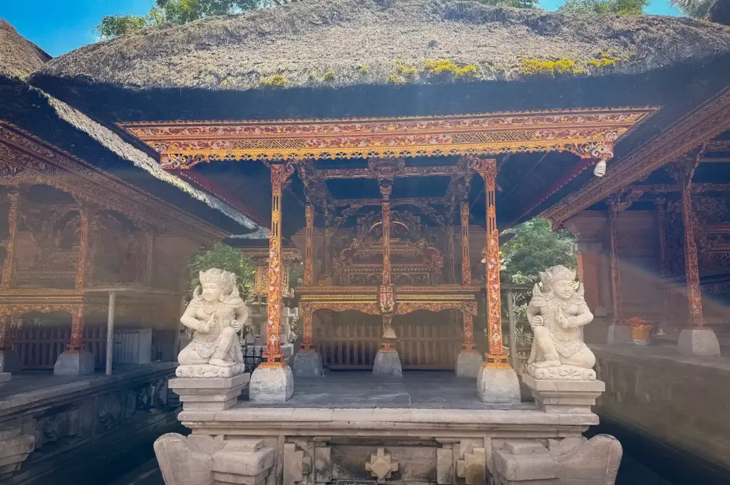 An architectural view of Pura Tirta Empul in Bali, featuring a traditional thatched-roof shrine adorned with intricate woodwork, colorful carvings, and stone guardian statues
