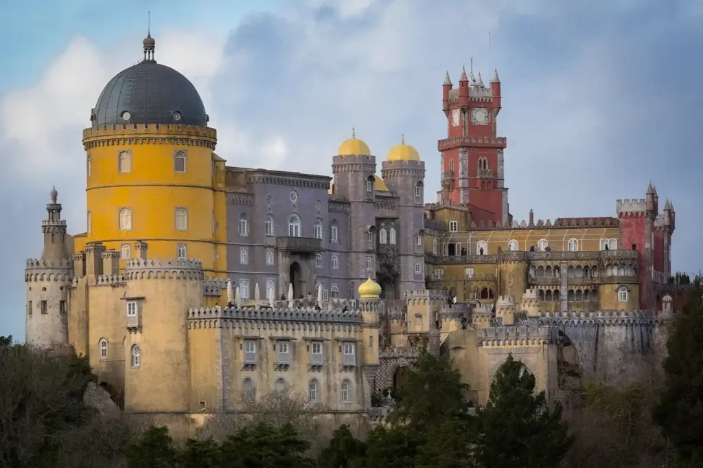 Exterior view of the colorful romanticist Pena Palace in Sintra, Portugal, featuring its distinct yellow and red towers, domes, and battlements set against a cloudy sky