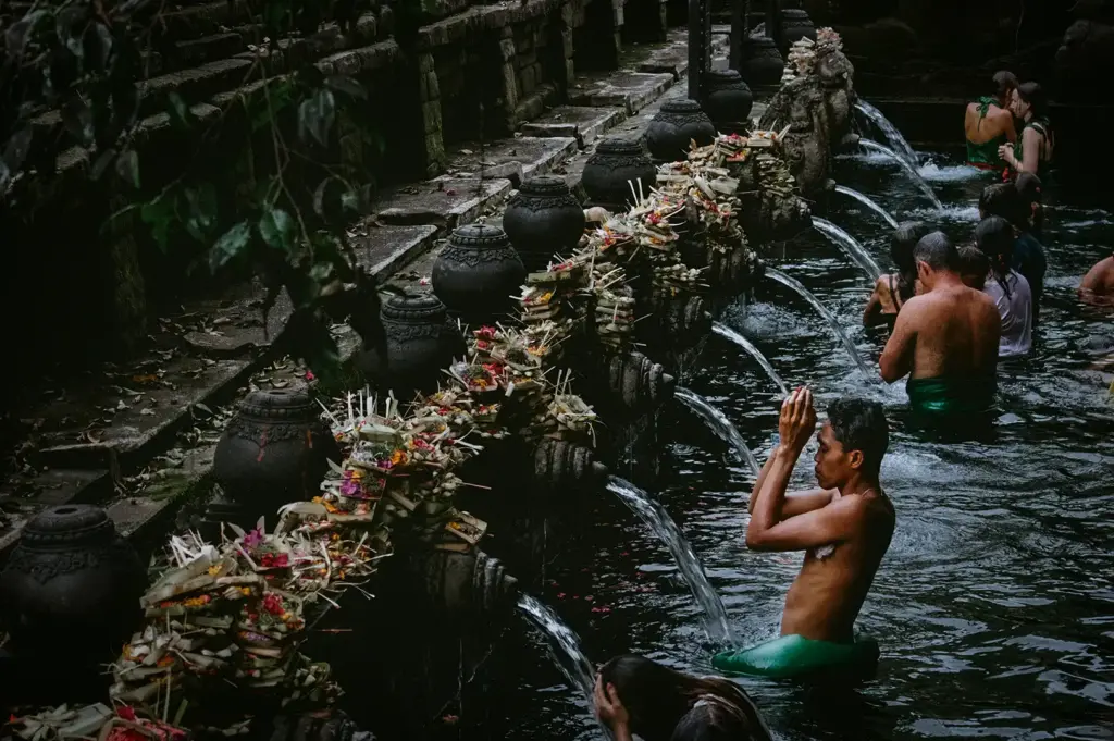 A traditional purification ritual at the sacred spring water pools of Pura Tirta Empul in Bali, featuring devotees offering prayers beneath flowing stone water spouts decorated with offerings.