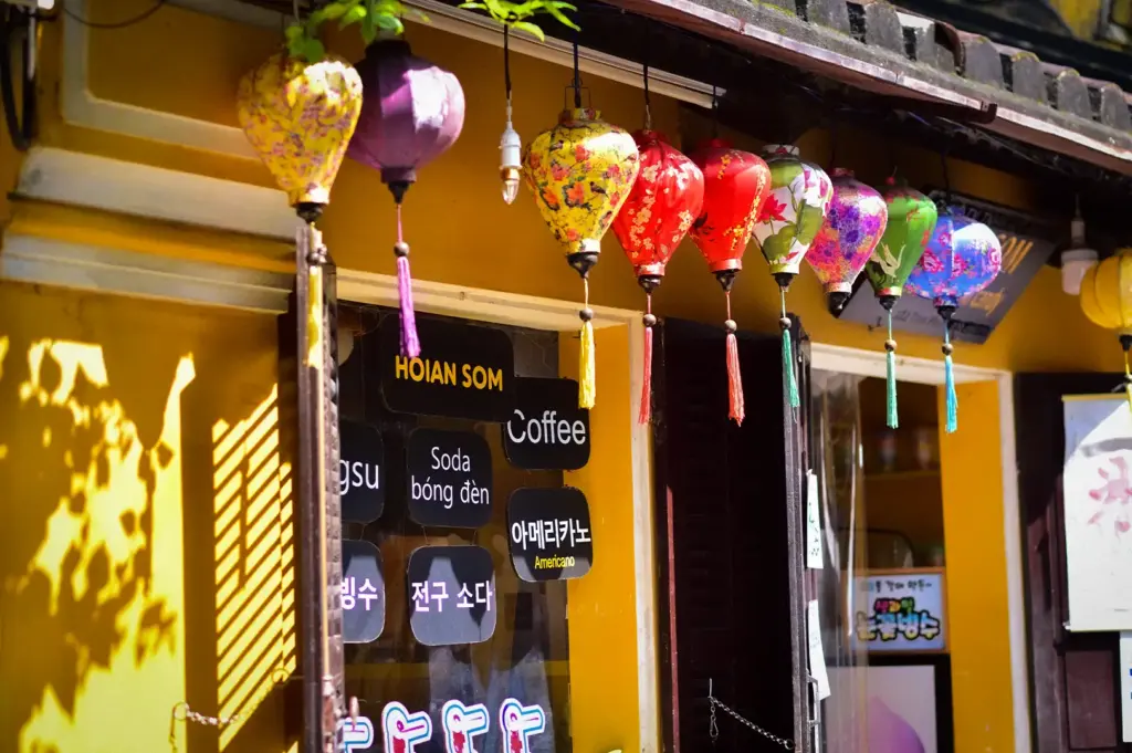 A row of colorful patterned silk lanterns hanging outside Hoian Som cafe with yellow walls and menu signage in Hoi An