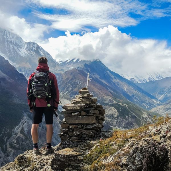 A solo traveler with a backpack stands triumphantly on a mountain peak, overlooking a vast landscape.